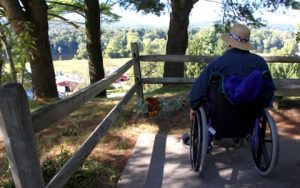 man sitting in wheelchair in front of fence overlooking trees, field and building