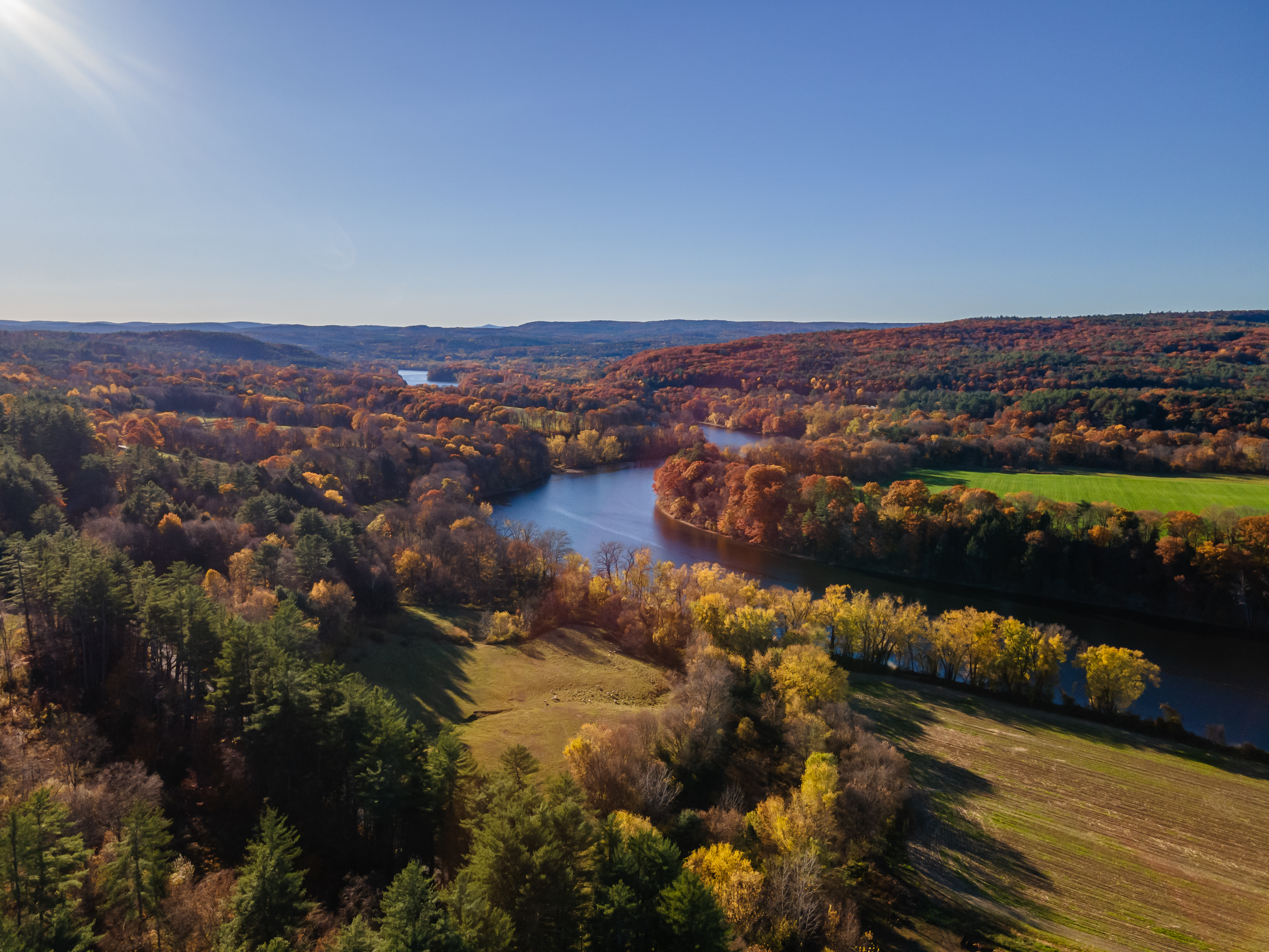 Aerial view of a winding river surrounded by colorful autumn trees, fields, and rolling hills under a clear blue sky.