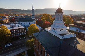 Aerial view of a small town with a prominent white cupola in the foreground, a church steeple, and tree-lined streets on a sunny day.