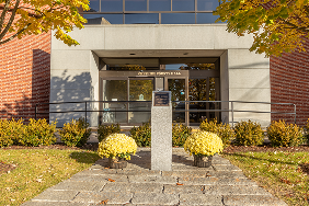Entrance to a building with a glass door, a central stone monument with a plaque, and yellow potted flowers on either side, surrounded by shrubs and autumn trees.
