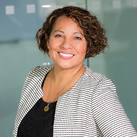 Woman with short curly hair wearing a striped blazer and black top, smiling and standing in front of a glass wall.