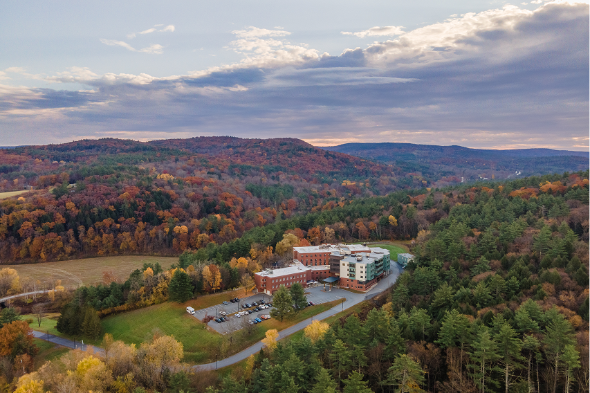 Aerial view of a large building complex surrounded by trees with autumn foliage and hills under a partly cloudy sky.