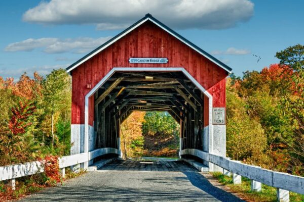 A red covered bridge with white trim spans a tree-lined road in autumn, with a sign indicating a 13-ton weight limit and clear skies above.
