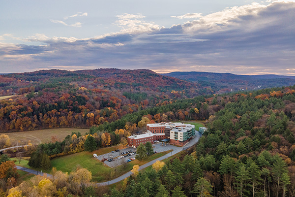 Aerial view of a large building complex surrounded by trees with autumn foliage, set in a hilly rural landscape under a partly cloudy sky.