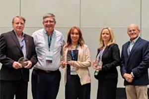 Five people stand side by side indoors; three hold glass awards, and all are dressed in business attire, posing for a group photo against a plain background.
